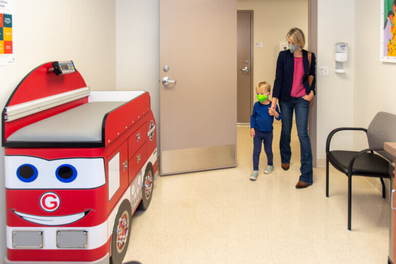A woman and child enter an exam room that features an exam bed decorated like a toy fire engine.