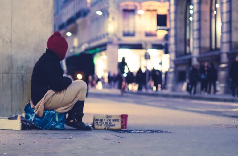 A person sits on a box with another box in front of him, asking for spare change.