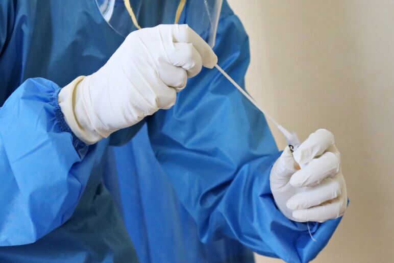 Photo of person in a medical gown and latex gloves, dipping a long nasal swab into COVID-19 testing solution